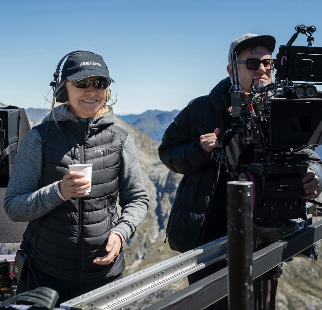 Director Charlotte Brändström, drink in hand, is seen with New Zealand scenery behind her and a camera operator on her left - on set for Amazon's Lord of the Rings tv show.