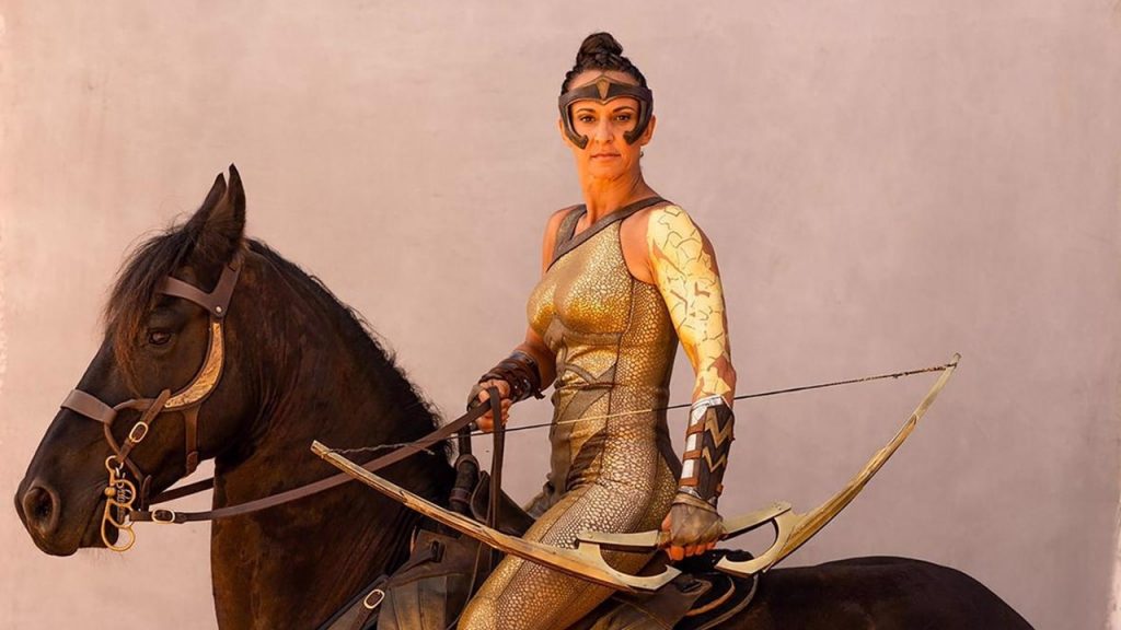 Stuntwoman Dayna Grant, as she appeared in Wonder Woman - mounted on dark brown horse, wearing a golden catsuit and with a golden bow in her left hand.