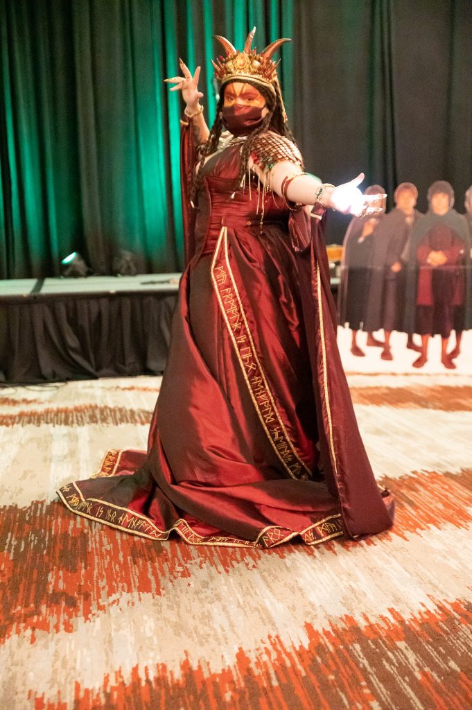 An amazing Smaug cosplay - a long, burgundy gown, with train edged with hand embroidered runes in gold. The dress includes scaled 'armour' epaulettes  in gold and burgundy, a flowing cloak, and  horned crown. The cosplayer has an illuminated Arkenstone in her hand, and long 'claw' fingernails. 