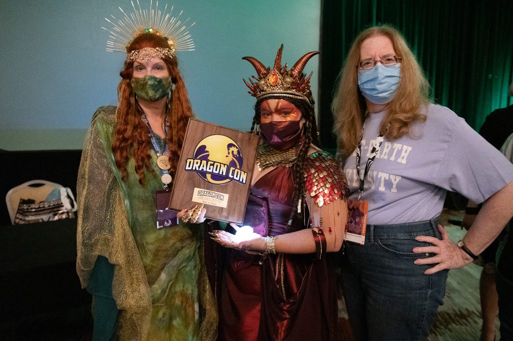 Best in Show winner in her Smaug gown is shown with her plaque award from DragonCon. On either side of her are costume contest judges Laura (L) and Constance (R). All are wearing masks over nose and mouth. 