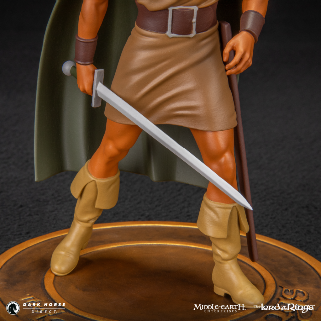Action figure of a fantasy warrior standing on a wooden base, wearing a tan tunic and cape, gripping a sword with a sheathed blade.
