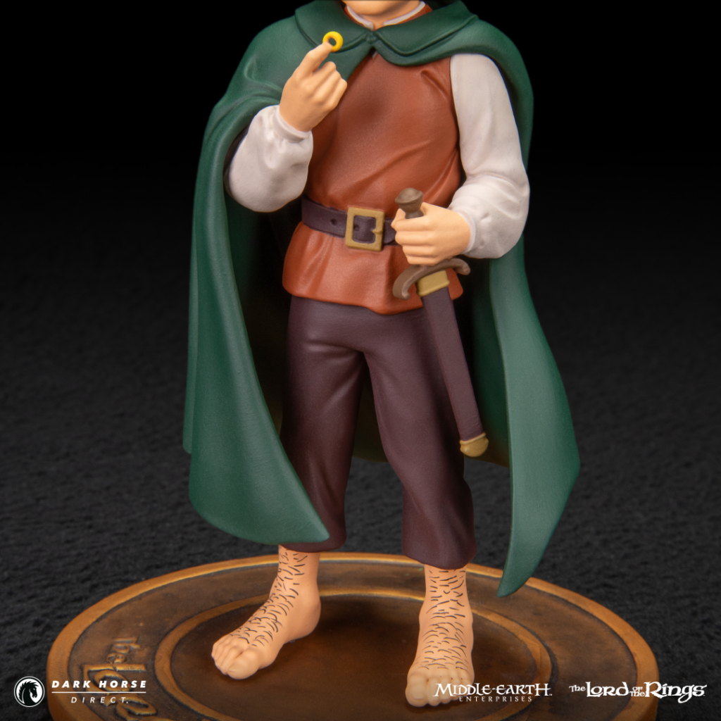 Action figure of a Hobbit with a green cape, brown tunic, sword at the belt, and bare feet, holding a small ring in one hand and standing on a circular base.