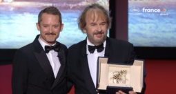 Two men in tuxedos smile on stage, holding an open award case with a palm-leaf trophy.