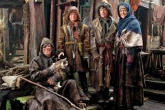 Man in a hooded cloak sits with a cat, while three children and a woman in fur robes stand beside him in a rustic wooden village setting.