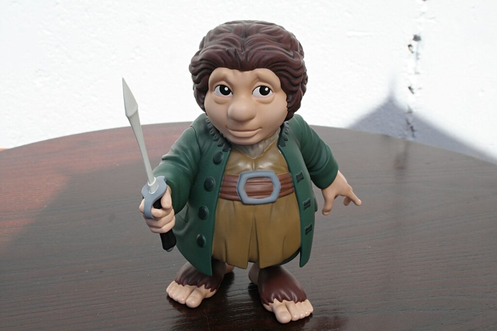 Plastic hobbit action figure in a green coat brandishing a small sword, standing on a wooden table