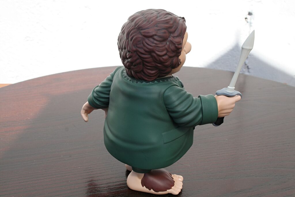 Back view of a plastic toy figure in a green outfit holding a sword on a wooden table surface.