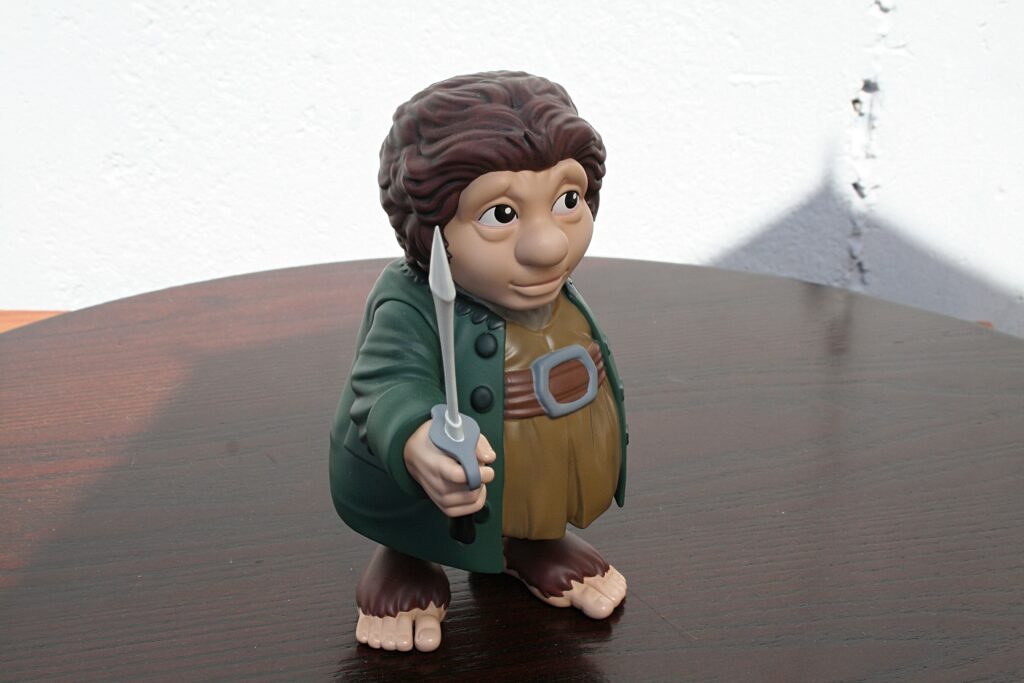 Toy figurine of a brown-haired adventurer in a green coat, holding a short sword, standing on a wooden table against a white wall.