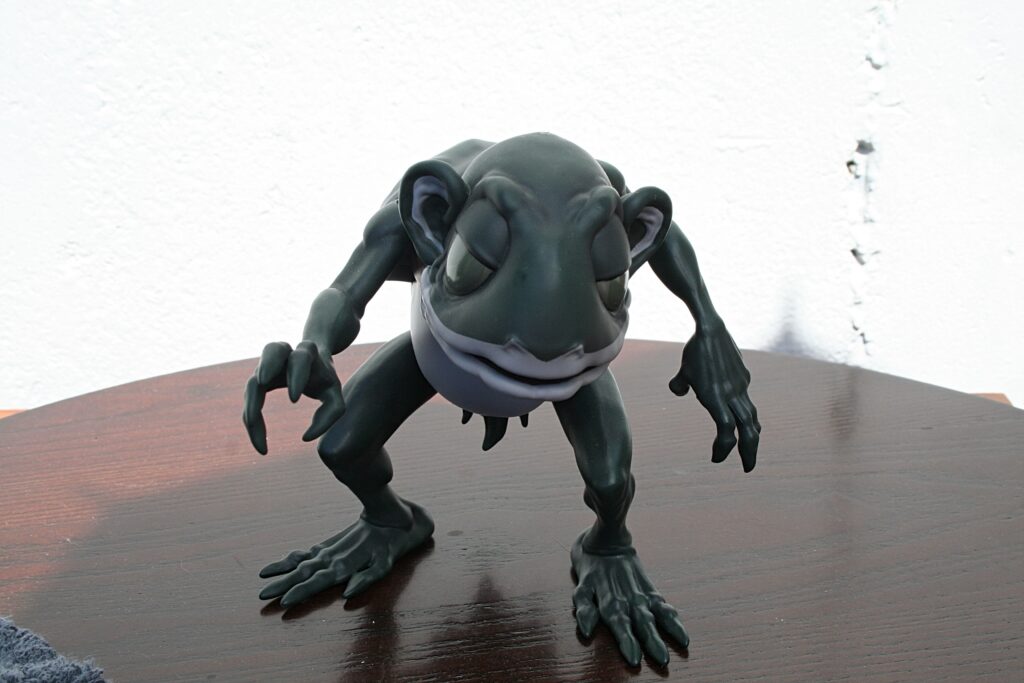 Gray alien-like sculpture crouching on a wooden table, reaching forward toward the viewer.