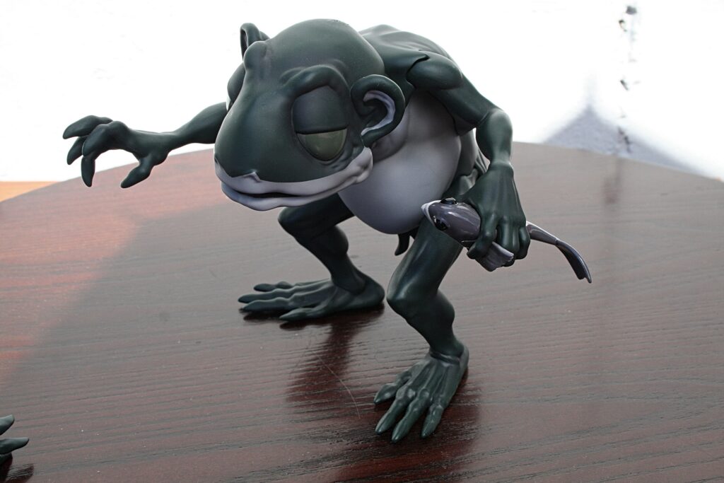 Dark green clay frog sculpture crouching on a polished wooden surface, one arm extended forward and fingers splayed.