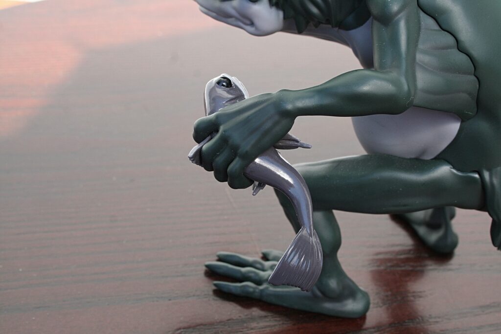 Close-up of a dark green statue's hands gripping a silver fish on a wooden floor surface.