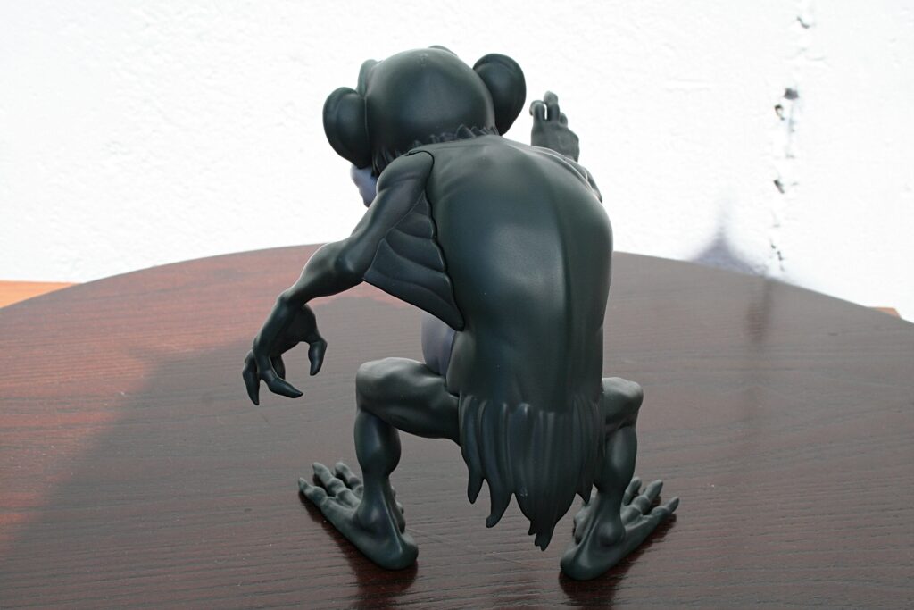 Dark sculpted creature crouching on a wooden table, viewed from behind, with a pale wall in the background.