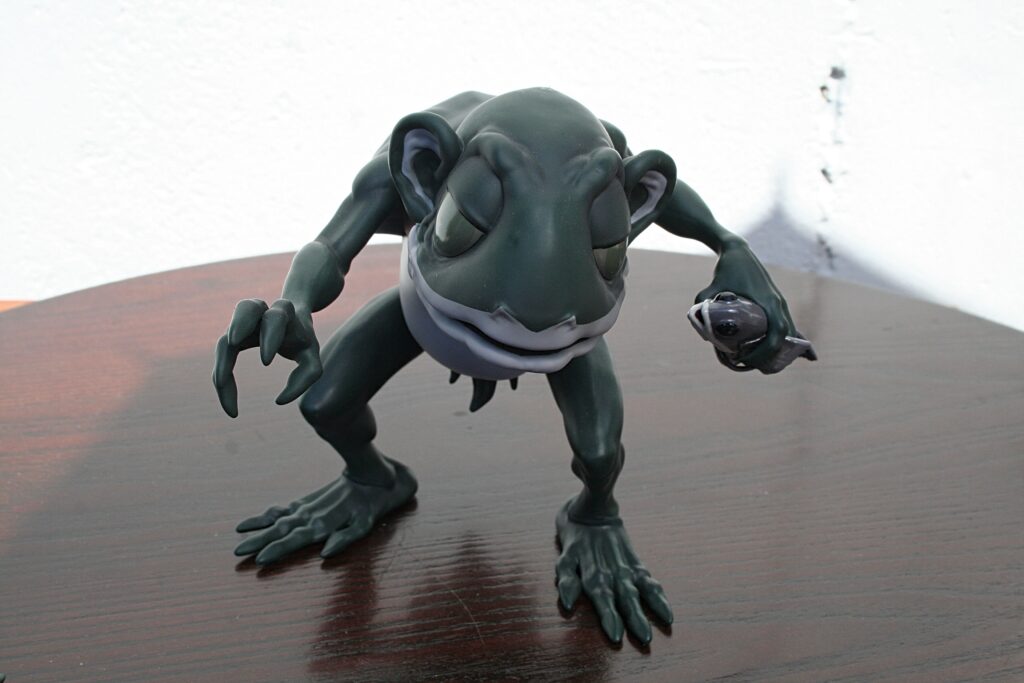 Front-facing view of a dark green clay alien/creature sculpture posed on a wooden table.