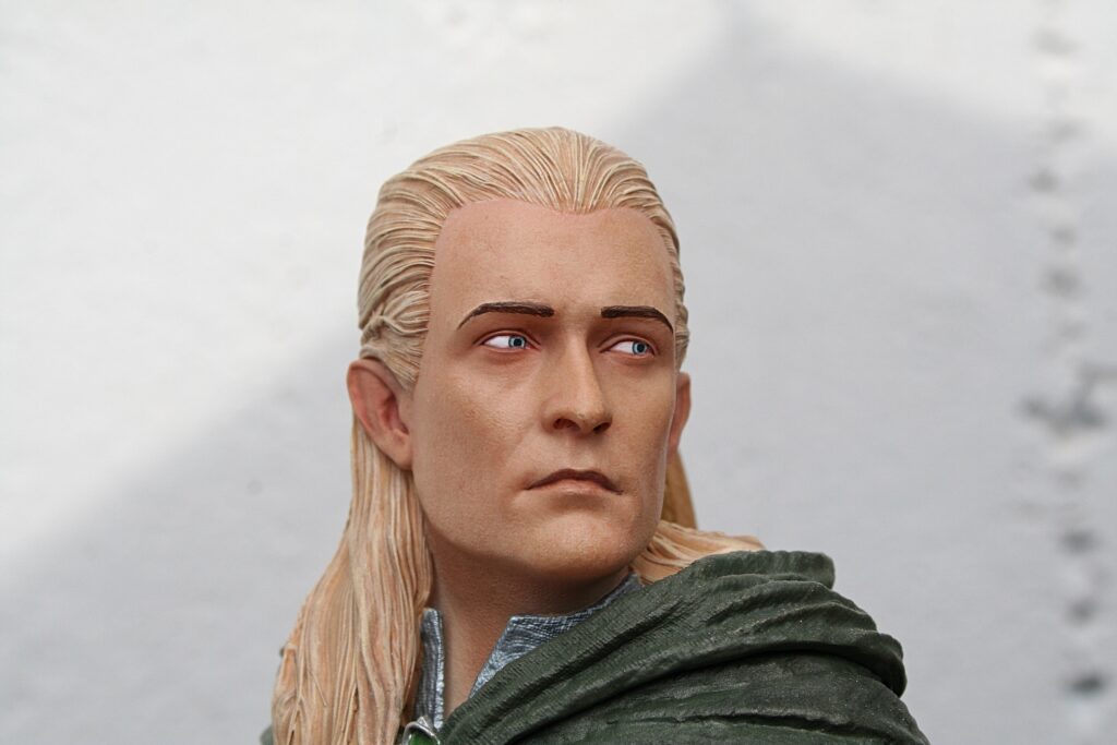 Close-up of a blond-haired action figure wearing a dark green cloak, looking to the right against a cloudy sky backdrop.