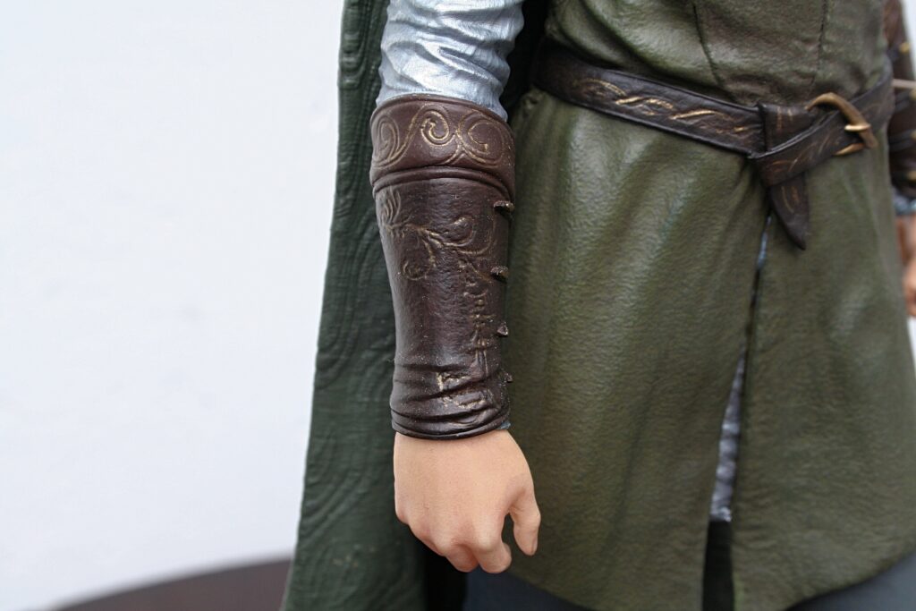 Close-up of a person wearing a brown patterned leather forearm bracer over a gray sleeve, with a green cloak and belt.