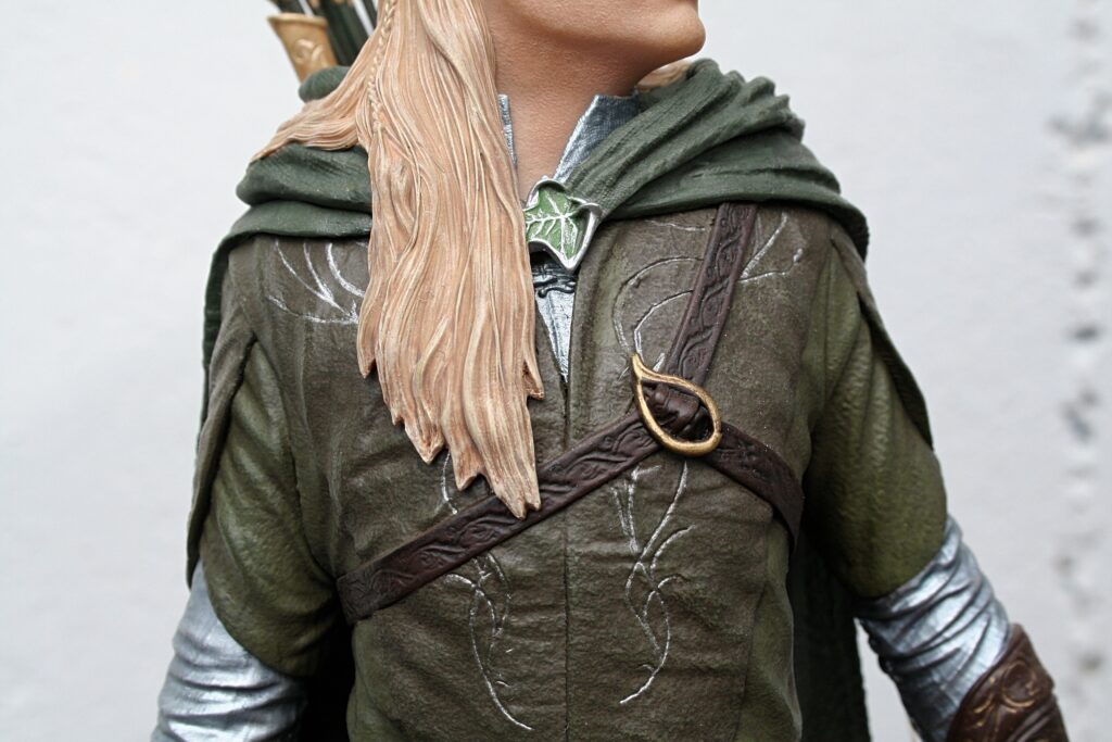 Cosplay close-up: person in a green medieval tunic with a brown strap, leaf-shaped brooch, and long light-brown hair.