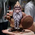 Plastic fantasy dwarf action figure held on an open hand, wielding a sword and round shield with a long gray beard and helmet.