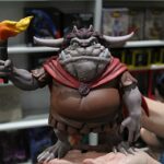 Toy horned demon figure holding a flame-tue torch atop a hand, with shelves in the background store setting.