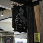 Dragon-themed banner with stylized text hanging from a dark wooden column in a convention hall, beside a yellow Lord of the Rings Fan Club poster featuring a QR code.