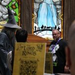 Cosplayer wearing a gray wizard costume and pointy hat at a convention booth, with colorful stained-glass panels in the background.