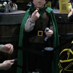 Cosplay character wearing a green cape and brown tunic, holding a ring up at a convention venue.