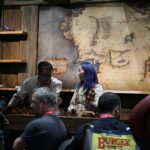 People stand at a wooden counter in front of a large antique map mural; shoppers in the foreground