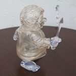 Clear glass figurine of a person holding a dagger, displayed on a dark round table.