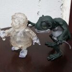 Clear plastic action figure with a raised sword stands on a dark wooden table while a green dragon figurine looms nearby.