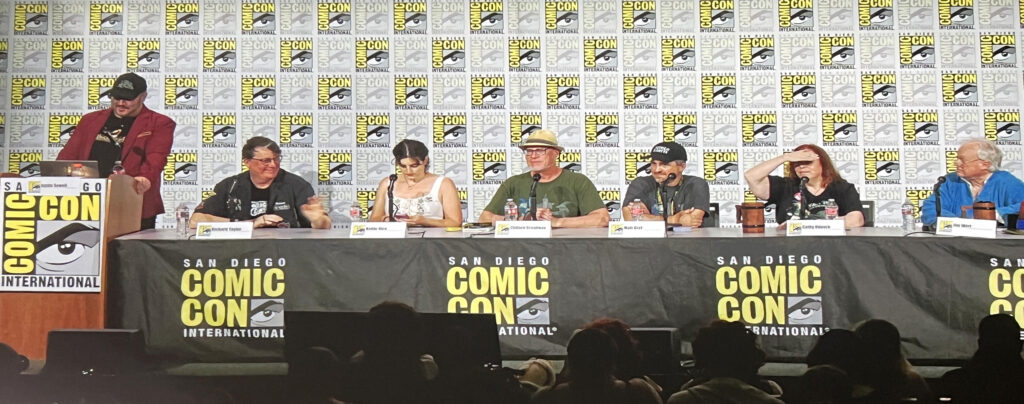 Panel of seven people seated at a long table on a Comic-Con stage, microphones in front of each, backdrop covered with Comic-Con logos.
