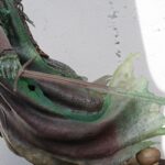 Close-up of a weathered statue hand with green patina gripping a rusted sword against a light gray wall.