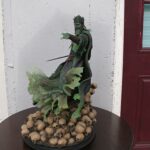 Statue of a green-clad king figure pointing forward, atop a mound of skulls on a round pedestal.