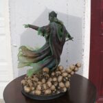 Green-cloaked king statue with a crown, arm extended as if pointing, atop a pile of skulls on a round table against a white wall and red door.