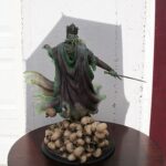 Statue of a crowned, robed king holding a sword, standing over a pile of skulls on a round pedestal.