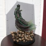 Green-robed crowned statue standing on a pile of skulls atop a round pedestal, against a white wall and a red door in the background.