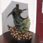 Green-robed crowned statue of a king, arm extended as if pointing, set on a dark round pedestal with a mound of walnuts around its base.