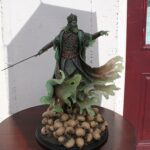 Green-clad king statue with crown, pointing forward, on a round table with a bed of walnuts underneath it.