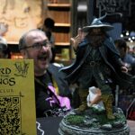 Actor-like wizard statue (Gandalf) on a rocky pedestal at a convention booth with 'The Lord of the Rings Fan Club' sign nearby.