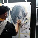 Artist wearing a cap and towel over his shoulder paints a large grayscale portrait with blue eyes on a taped board at an art exhibit