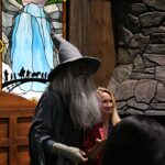 People in wizard costumes stand near a stained-glass panel depicting a waterfall in a themed setting.