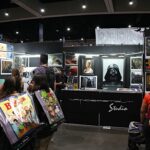 Comic convention booth with framed artwork on dark panels and people browsing the displays, some carrying bags and backpacks.