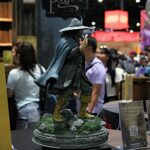 Life-sized fantasy statue of a cloaked, goblin-like figure standing on a rocky pedestal at a convention booth with people and colorful banners in the background.