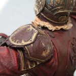 Close-up of red, embossed armor with gold trim on shoulder/back plates, showing intricate decorative details.