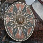 Close-up of a round, ornate shield with a sunburst metal design mounted on armored leather armor of a fantasy warrior.