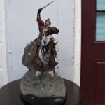 Statue of a medieval knight in ornate armor, sword raised high, mounted on a rocky pedestal.
