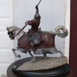 Armored knight statue atop a charging horse, sword raised high, on a textured pedestal beside a white door and a red-framed window.