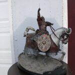 Knight in armor on a rearing horse statue, sword raised, with decorative shield and armor plates on a rocky pedestal