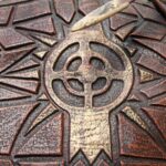 Close-up of a carved wooden panel with a central skull-like motif and geometric, sunburst patterns in reddish-brown tones