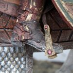 Close-up of a decorative armored boot with red leather, laces and gold filigree next to an ornate shield with pink gems.