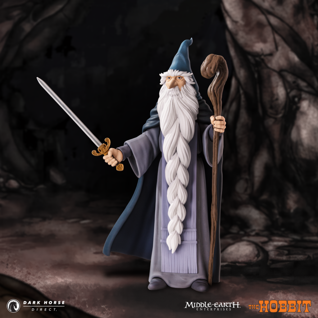 Action figure of Gandalf from The Hobbit, wearing a blue cloak, holding a sword in one hand and a staff in the other in a dark cave setting.