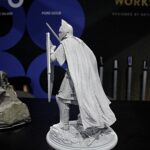 Gray armored knight figurine with cape, shield, and spear standing on a rocky pedestal in a display scene