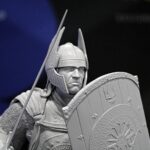Gray knight figurine in armor, holding a spear and a heavily engraved shield close up.